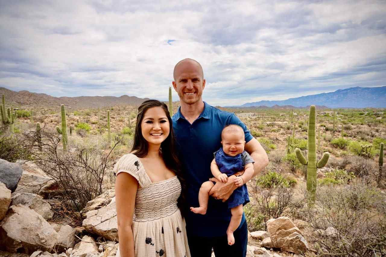Family portrait in Arizona desert with cacti, featuring a smiling woman in a floral dress, a man in a blue shirt holding a happy baby in denim, showcasing the region's natural beauty and warmth.